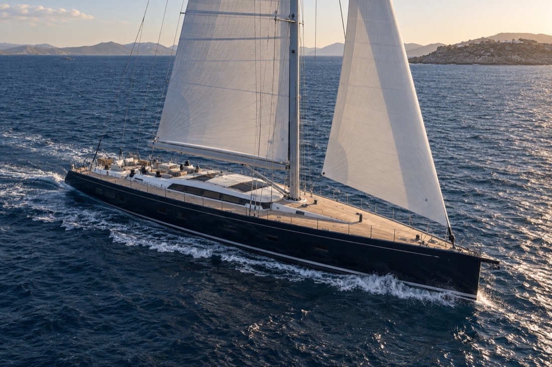 Long blue-hulled sailing monohull under full sail beside a Mediterranean coastline